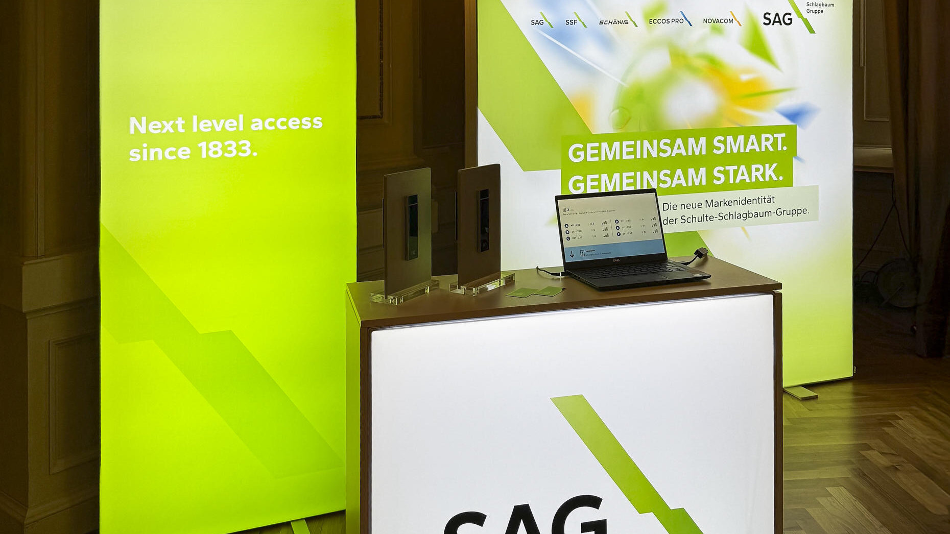 Foto vom Markenauftritt der SAG bei der Hauptversammlung 2025. Weiße Theke mit schwarzer SAG Aufschrift und Grünen stilistischen Schlagbaum (SAG Logo). Im Hintergrund eine schmale grüne Messewand und eine Quadratische Messe Wand mit dem Slogan Gemeinsam smart, Gemeinsam Stark