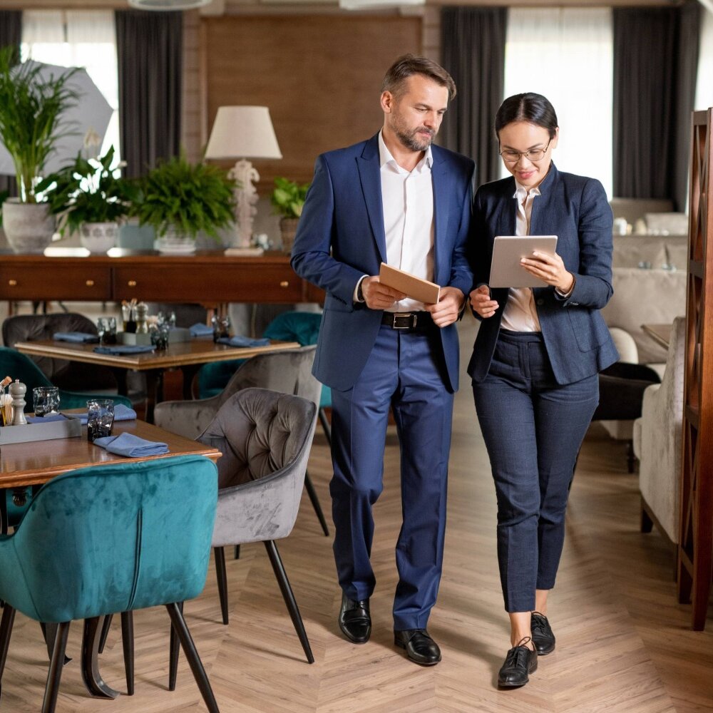 Ein Mann und eine Frau im Anzug stehen in einem modern eingerichtetem Restaurant und schauen auf einen kleinen Bildschirm