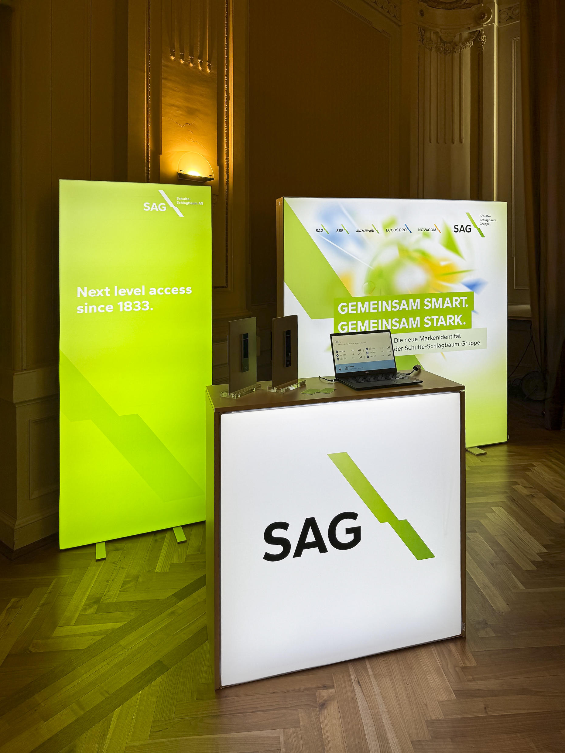 Foto vom Markenauftritt der SAG bei der Hauptversammlung 2025. Weiße Theke mit schwarzer SAG Aufschrift und Grünen stilistischen Schlagbaum (SAG Logo). Im Hintergrund eine schmale grüne Messewand und eine Quadratische Messe Wand mit dem Slogan Gemeinsam smart, Gemeinsam Stark