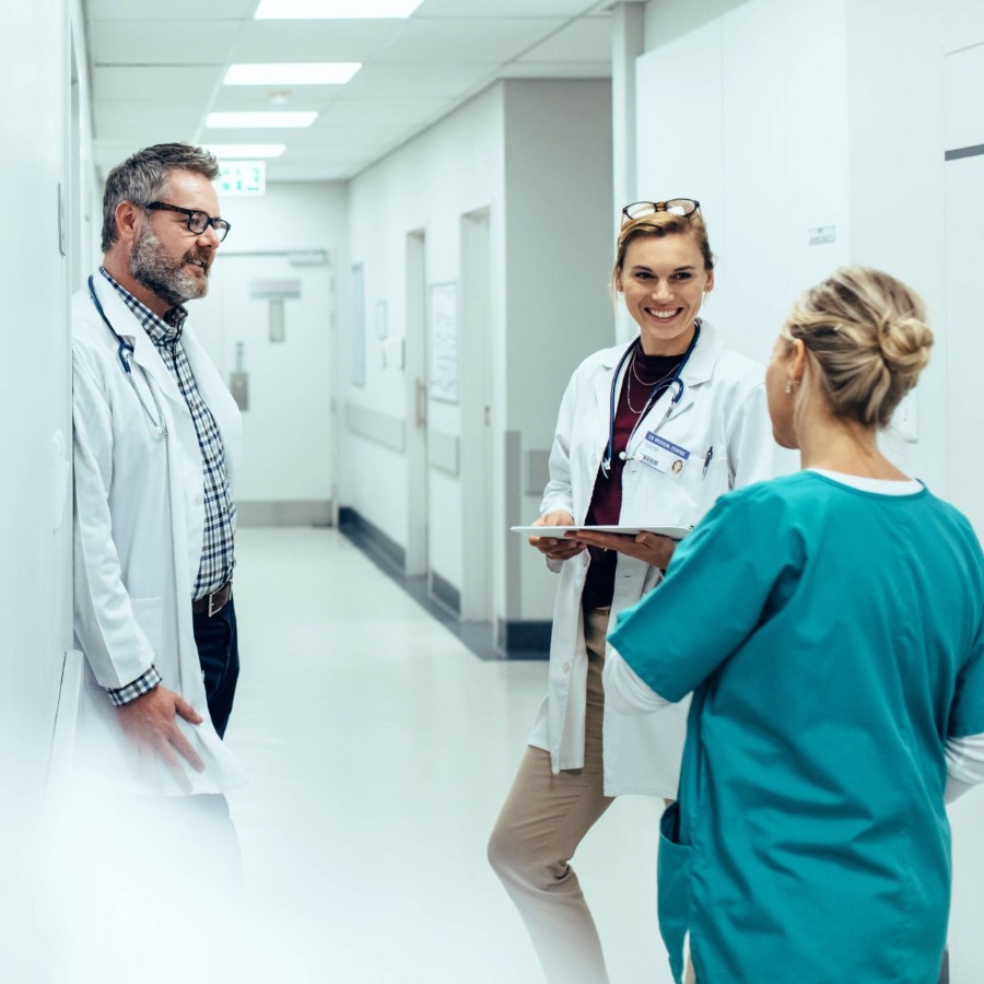 Ein Arzt und eine Ärztin unterhalten sich freundlich mit einer Krankenschwester, im Flur eines Krankenhauses